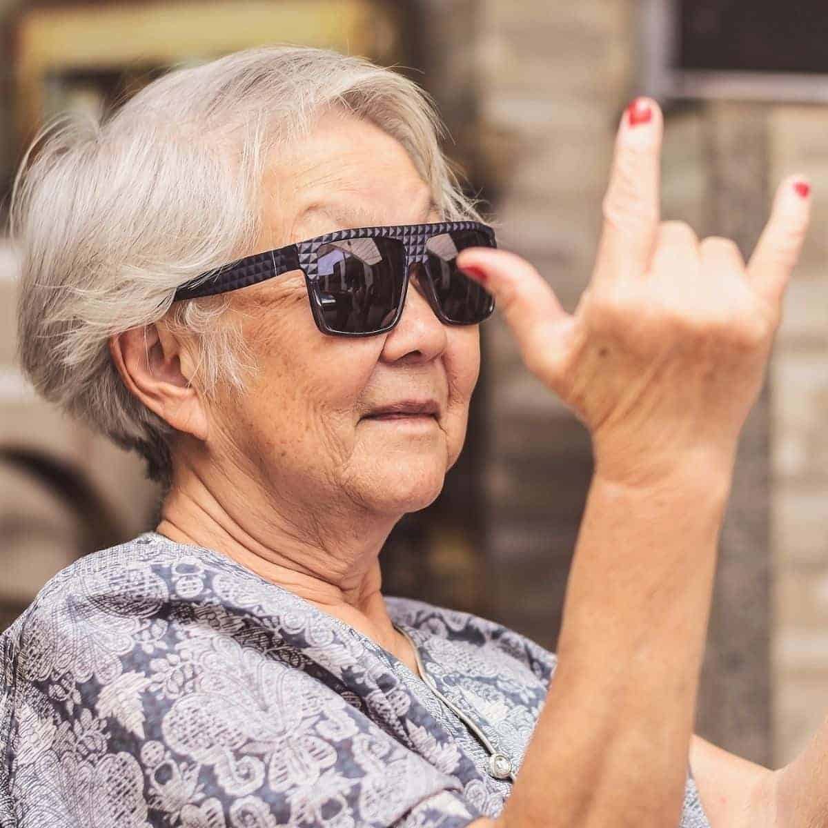Old woman sitting down and wearing sunglasses with her hands up.